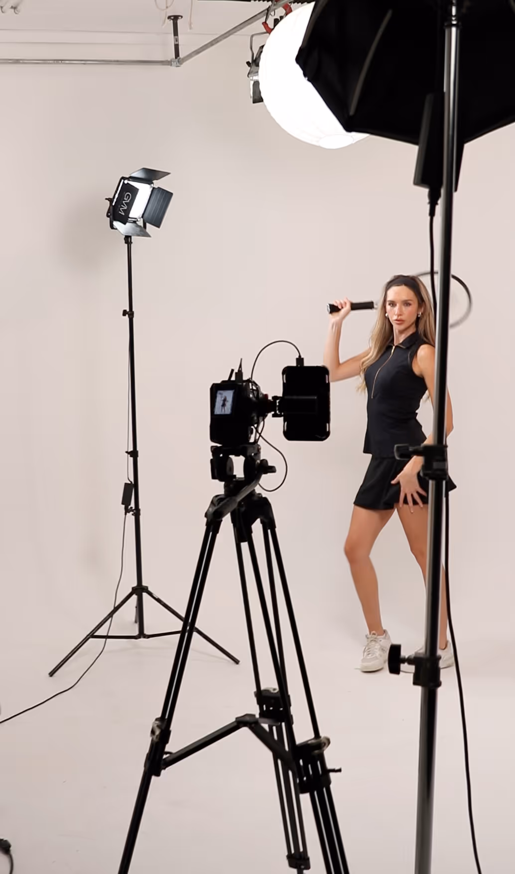Woman in black sleeveless top and skirt posing with a tennis racket in a photo studio with lighting and camera equipment.